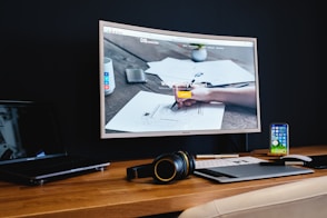 A modern workspace features a large curved monitor displaying a web agency page, surrounded by various items including a laptop, a smartphone, a wireless keyboard, headphones, and a stylus pad. The setup is neatly arranged on a wooden desk against a dark wall, creating a sleek and professional look.
