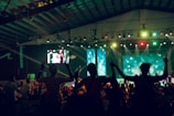 A large indoor event with a stage and a crowd of people. The audience appears to be enjoying a performance as they raise their hands in celebration. Colorful lights illuminate the venue, creating a vibrant atmosphere. Two large screens are visible in the background, displaying various visuals.