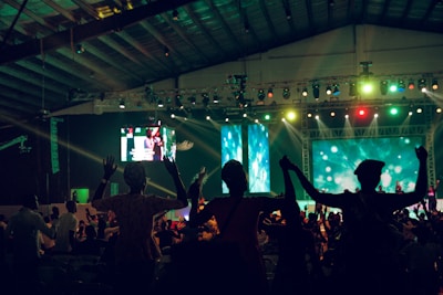 A large indoor event with a stage and a crowd of people. The audience appears to be enjoying a performance as they raise their hands in celebration. Colorful lights illuminate the venue, creating a vibrant atmosphere. Two large screens are visible in the background, displaying various visuals.