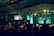 A large indoor event with a stage and a crowd of people. The audience appears to be enjoying a performance as they raise their hands in celebration. Colorful lights illuminate the venue, creating a vibrant atmosphere. Two large screens are visible in the background, displaying various visuals.