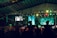 A large indoor event with a stage and a crowd of people. The audience appears to be enjoying a performance as they raise their hands in celebration. Colorful lights illuminate the venue, creating a vibrant atmosphere. Two large screens are visible in the background, displaying various visuals.
