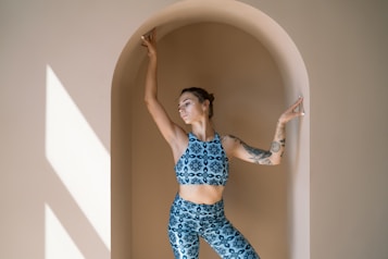A woman with tattoos is striking a graceful pose against an archway bathed in soft natural light. She is wearing a matching blue patterned sports bra and leggings set. Her arms are elegantly extended, and she looks calm and focused.