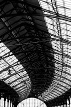 A large, intricate metal and glass structure features a series of curved steel beams and panels. The architecture showcases a complex pattern of lines and geometric shapes, emphasizing industrial design elements.