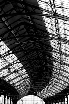 A large, intricate metal and glass structure features a series of curved steel beams and panels. The architecture showcases a complex pattern of lines and geometric shapes, emphasizing industrial design elements.