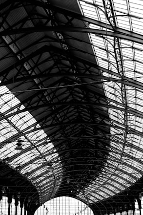 A large, intricate metal and glass structure features a series of curved steel beams and panels. The architecture showcases a complex pattern of lines and geometric shapes, emphasizing industrial design elements.