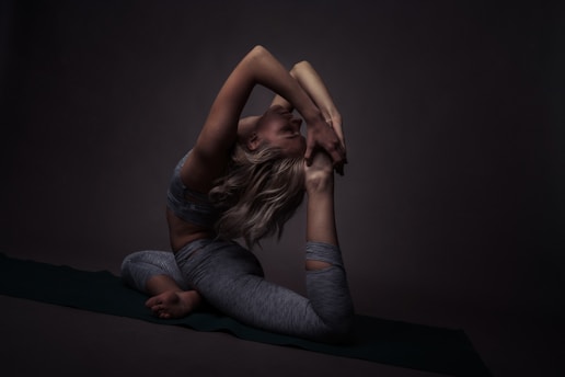 A person in a challenging yoga pose, balancing gracefully on a mat. The surrounding environment is dimly lit, creating deep shadows that highlight the contours and muscles. The focus is on the flexibility and strength of the pose.
