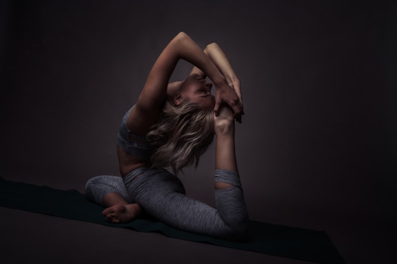 A person in a challenging yoga pose, balancing gracefully on a mat. The surrounding environment is dimly lit, creating deep shadows that highlight the contours and muscles. The focus is on the flexibility and strength of the pose.