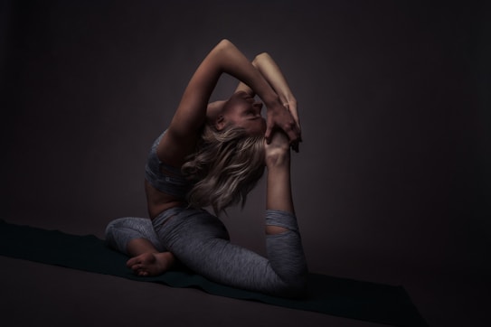 A person in a challenging yoga pose, balancing gracefully on a mat. The surrounding environment is dimly lit, creating deep shadows that highlight the contours and muscles. The focus is on the flexibility and strength of the pose.