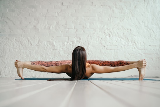 A person is engaging in a yoga pose, stretching their legs apart while lying on a mat with their arms extended, holding their feet.