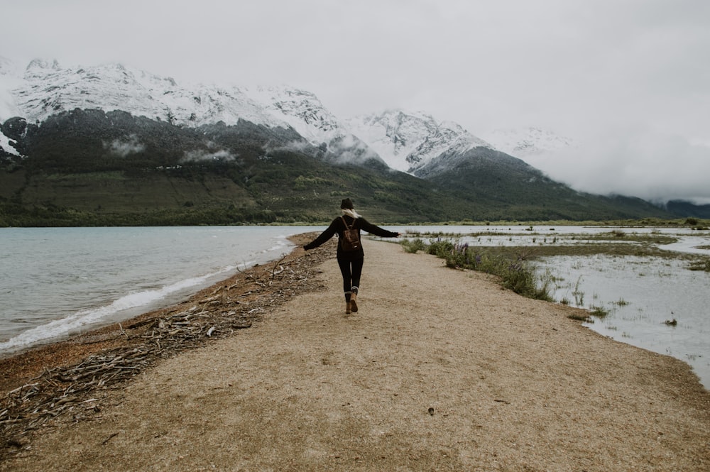 Mujer con camisa negra de manga larga y pantalones negros caminando por un camino de tierra marrón cerca del lago