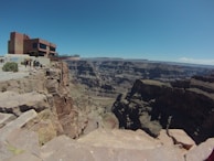 A modern building perched on the edge of a vast canyon, with tourists walking along the cliff. The deep, rugged canyon stretches into the distance, showcasing layers of rock formations under a clear blue sky.