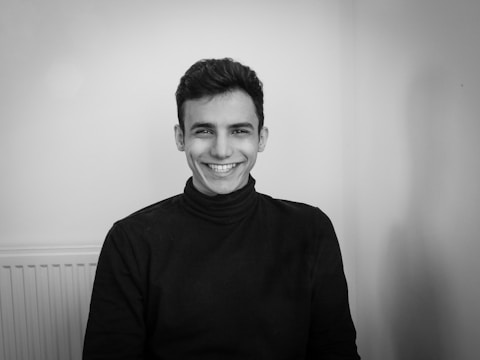 A black and white portrait of a person smiling warmly while seated in front of a plain background. Wearing a dark turtleneck sweater, the person exudes a friendly and approachable demeanor.