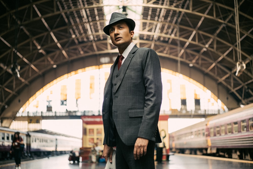 man in black suit jacket and black hat standing near bridge during daytime