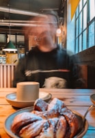 A person sits at a wooden table inside a dimly lit café with an industrial aesthetic. Their face is blurred due to motion, giving a sense of movement or obscured identity. On the table, a cappuccino in a beige cup and a plate with a pastry dusted with powdered sugar are prominently visible. In the background, a green hanging lamp, windows with natural light, and some wall decor can be seen.