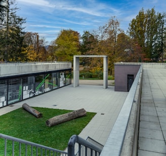 An outdoor area featuring a modern architectural design with a flat, paved surface and a patch of green grass. Two large wooden logs are placed on the grass. The surrounding buildings have large glass windows and there are trees with autumn foliage in the background.