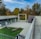 An outdoor area featuring a modern architectural design with a flat, paved surface and a patch of green grass. Two large wooden logs are placed on the grass. The surrounding buildings have large glass windows and there are trees with autumn foliage in the background.