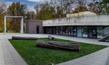 A modern building with a concrete facade and large glass windows is surrounded by a tidy garden area. The structure includes a flat roof and an overhang above the entrance. In the foreground, several large logs are placed on a patch of green grass, creating a landscaped area within a plaza paved with large stone tiles. Tall trees with autumn foliage are visible in the background, contributing to the serene, natural setting.