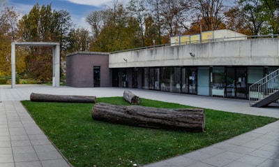 A modern building with a concrete facade and large glass windows is surrounded by a tidy garden area. The structure includes a flat roof and an overhang above the entrance. In the foreground, several large logs are placed on a patch of green grass, creating a landscaped area within a plaza paved with large stone tiles. Tall trees with autumn foliage are visible in the background, contributing to the serene, natural setting.