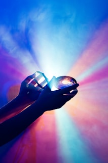 Hands are holding a crystal or prism, casting vibrant and colorful light rays. The light refracts into a spectrum of colors, producing a visually striking and ethereal effect against a softly blurred background.