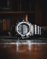 A rugged digital wristwatch with a robust black frame and various features including altimeter, barometer, and multiple sensors. It is positioned on a reflective surface with a blurred background, suggesting an indoor setting.