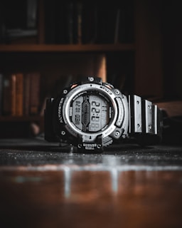 A rugged digital wristwatch with a robust black frame and various features including altimeter, barometer, and multiple sensors. It is positioned on a reflective surface with a blurred background, suggesting an indoor setting.