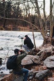 Two photographers are near a flowing river, surrounded by large rocks and bare trees. One is kneeling, setting up his camera on a tripod, while the other is sitting and photographing the scene. The environment is natural and serene, with a backdrop of a calm forest and cascading water.