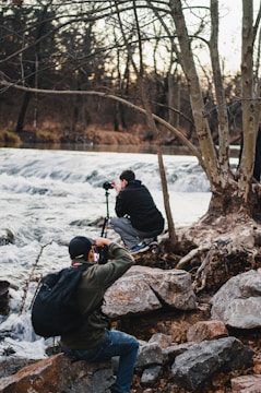 Two photographers are near a flowing river, surrounded by large rocks and bare trees. One is kneeling, setting up his camera on a tripod, while the other is sitting and photographing the scene. The environment is natural and serene, with a backdrop of a calm forest and cascading water.