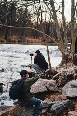 Two photographers are near a flowing river, surrounded by large rocks and bare trees. One is kneeling, setting up his camera on a tripod, while the other is sitting and photographing the scene. The environment is natural and serene, with a backdrop of a calm forest and cascading water.