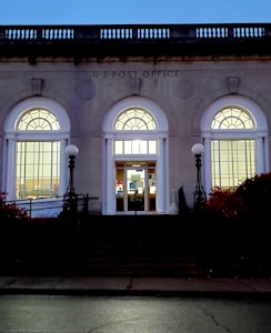 An elegant building facade with three large arched windows, each illuminated from the inside. The structure is labeled as a U.S. Post Office. Classic street lamps with spherical bulbs flank the entrance, and there are steps leading up to the doorway.