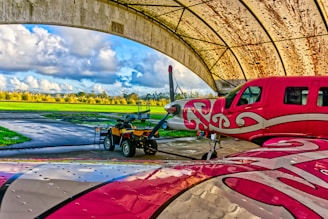 A brightly painted red and white airplane with swirling patterns is parked under a large archway hangar in an airfield. Next to it, a small yellow and black towing vehicle is visible. The background features a lush green field under a partly cloudy sky.