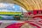 A brightly painted red and white airplane with swirling patterns is parked under a large archway hangar in an airfield. Next to it, a small yellow and black towing vehicle is visible. The background features a lush green field under a partly cloudy sky.