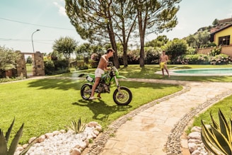 A sunny garden setting with a stone path and lush green lawn is depicted. A person is standing on the grass with a green motocross bike, wearing shorts. Another person in swimwear stands by the swimming pool, surrounded by flowers and trees. The scene conveys a summery and leisure-filled atmosphere.