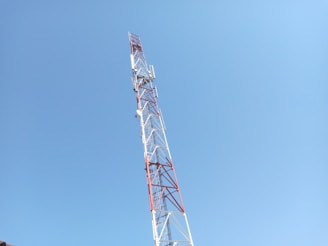 A tall communication tower with red and white metal structures extends upwards against a clear blue sky. Several antennas and satellite dishes are attached to the tower at various points.