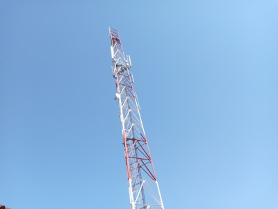 A tall communication tower with red and white metal structures extends upwards against a clear blue sky. Several antennas and satellite dishes are attached to the tower at various points.