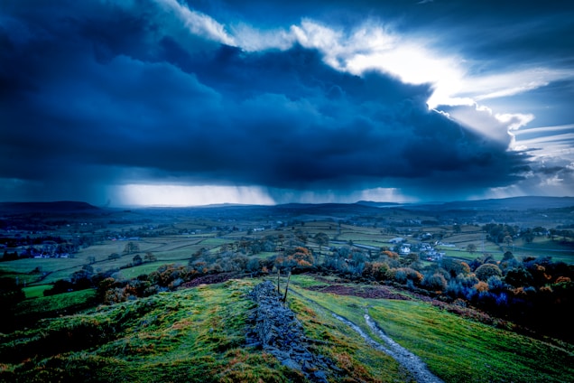 A dramatic landscape with a large, dark storm cloud dominating the sky, casting shadows over the vast, green fields below. The terrain is gently rolling with a mixture of grass and small trees, divided by stone walls. The horizon is dotted with small houses and hills, with the sun breaking through the clouds creating rays of light.