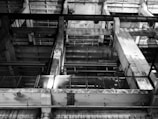 A black and white image of an industrial interior featuring large concrete structures and metal beams. The design has a grid-like pattern with visible pipes and railings, creating an intricate geometric composition.