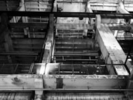 A black and white image of an industrial interior featuring large concrete structures and metal beams. The design has a grid-like pattern with visible pipes and railings, creating an intricate geometric composition.
