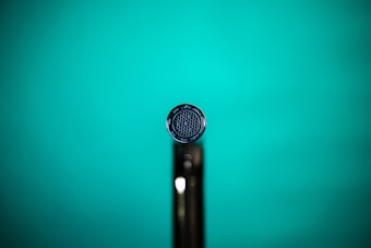 A close-up shot of a microphone head with a detailed grill pattern against a vibrant teal background. The image focuses on the front view of the microphone, capturing the intricate mesh design at the center.