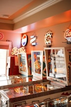 A room with several vintage pinball machines arranged in a row. Neon signs for various brands including Pepsi Cola are affixed to the wall above the machines. The ambiance is warm and nostalgic with a mix of bright and soft lighting.