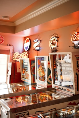 A room with several vintage pinball machines arranged in a row. Neon signs for various brands including Pepsi Cola are affixed to the wall above the machines. The ambiance is warm and nostalgic with a mix of bright and soft lighting.