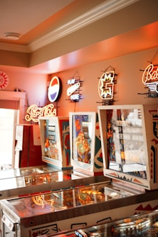 A room with several vintage pinball machines arranged in a row. Neon signs for various brands including Pepsi Cola are affixed to the wall above the machines. The ambiance is warm and nostalgic with a mix of bright and soft lighting.