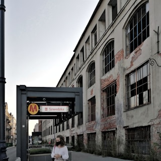 A woman wearing a mask stands near a metro sign for Szwedzka station, in front of an old, deteriorating building with large windows and exposed brickwork.