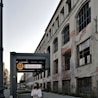 A woman wearing a mask stands near a metro sign for Szwedzka station, in front of an old, deteriorating building with large windows and exposed brickwork.