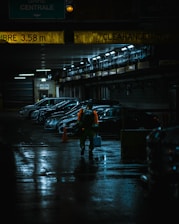 A dimly lit parking garage with several parked cars lined up against a wall. The ceiling has yellow clearance signs, and the floor is wet, reflecting light. A person wearing a bright orange safety vest walks towards the camera, carrying a blue container and a backpack.