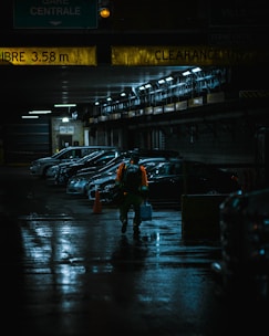 A dimly lit parking garage with several parked cars lined up against a wall. The ceiling has yellow clearance signs, and the floor is wet, reflecting light. A person wearing a bright orange safety vest walks towards the camera, carrying a blue container and a backpack.