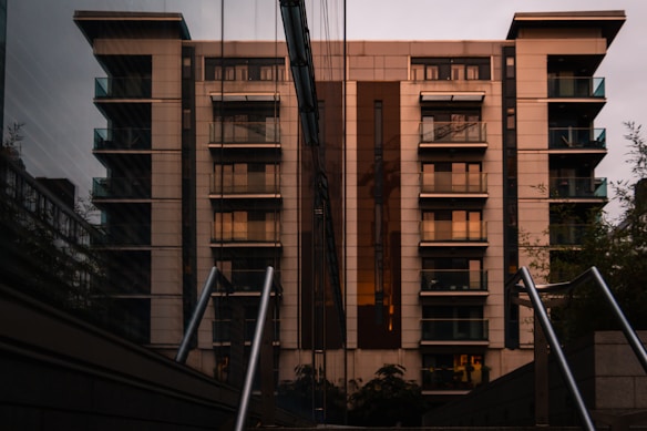 A modern apartment building with multiple balconies stands tall against a cloudy sky. The structure features clean lines and a mix of glass and stone materials, reflecting the minimalistic architectural style. The image also captures a reflection of the building on an adjacent glass surface, adding depth and symmetry.
