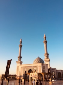 A grand mosque with two tall minarets and a large central dome is situated under a clear blue sky. The architecture features intricate designs, and there are several people walking in the foreground. A black flag with white calligraphy stands prominently to the left.