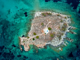 An aerial view of a small, rocky island surrounded by clear turquoise water. The island features sparse vegetation, mainly shrubs and small trees. A lone structure with a blue roof and white exterior stands prominently in the center, surrounded by a dirt path leading from the shore.
