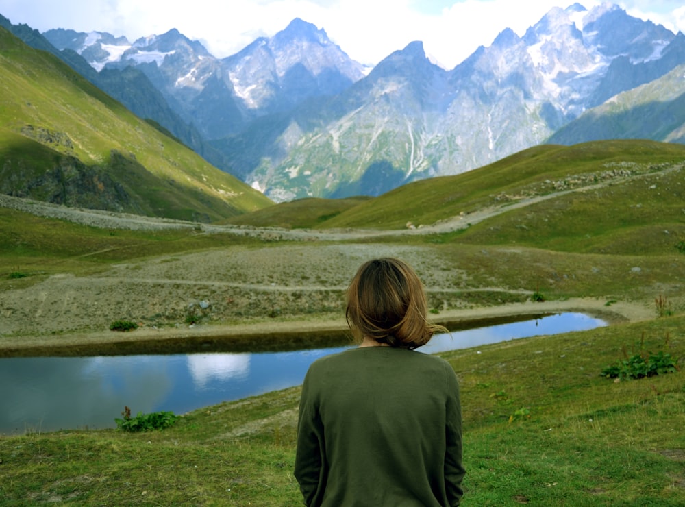 woman in green shirt standing on green grass field near lake and mountains during daytime