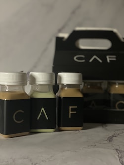 Three small bottles with white caps and dark labels are arranged in front of a black carrier box. The letters 'C', 'A', and 'F' are visible on the bottles. The background is a marble-like surface.